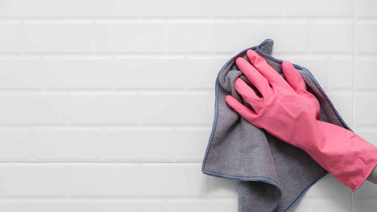 Mano con guante rosa limpiando azulejos blancos con un paño de microfibra